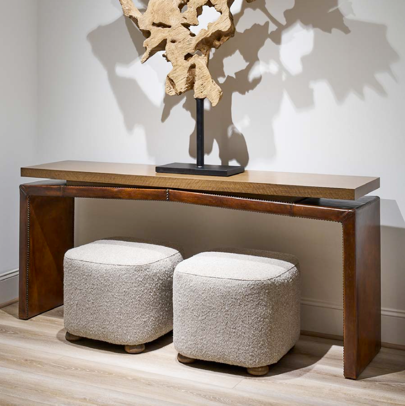Wood and leather console table with two upholstered stools and a piece of abstract art on a white wall.
